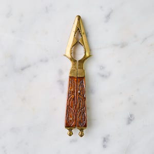 May include: A vintage nutcracker with a brass head and intricately carved wooden handles. The handles are a rich brown color with a detailed leaf pattern. The nutcracker is designed with a scissor-like mechanism for cracking nuts.