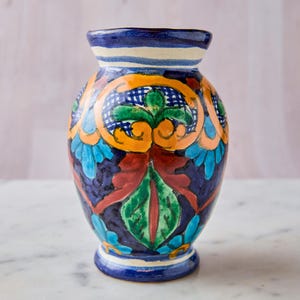 May include: A hand-painted ceramic vase with a dark blue base and colorful floral designs. The vase features orange, green, red, and blue accents. The rim is blue with white stripes. A decorative piece for the home.