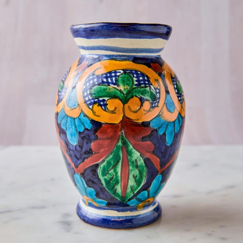 May include: A hand-painted ceramic vase with a dark blue base and colorful floral designs. The vase features orange, green, red, and blue accents. The rim is blue with white stripes. A decorative piece for the home.