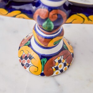 May include: A colorful, ceramic cake stand with a detailed, hand-painted design. The stand features a round base with a pattern of yellow, red, and blue floral motifs. The stem and top are also decorated with intricate patterns and vibrant colors.