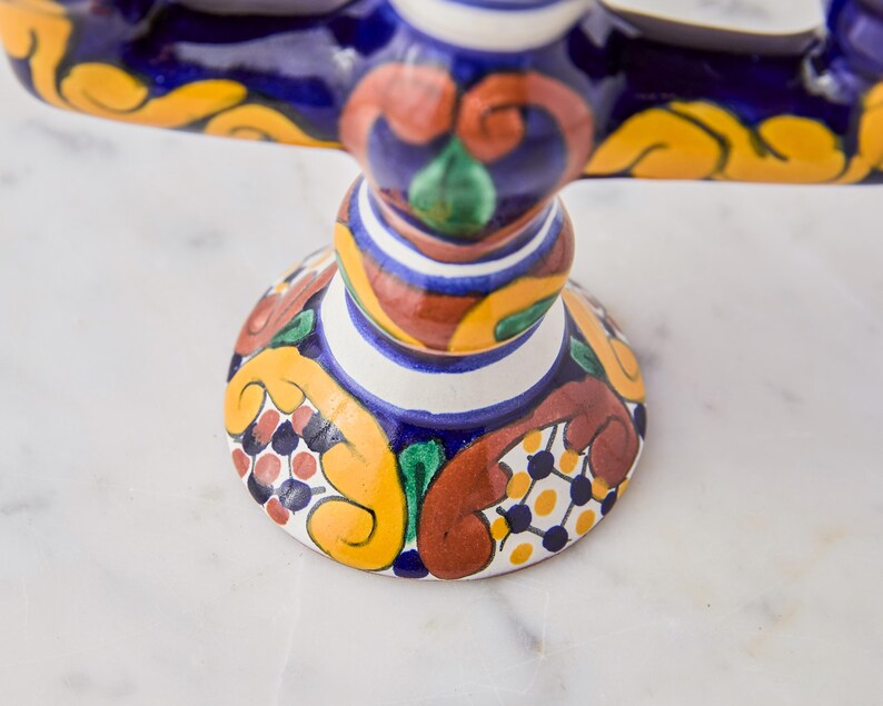 May include: A colorful, ceramic cake stand with a detailed, hand-painted design. The stand features a round base with a pattern of yellow, red, and blue floral motifs. The stem and top are also decorated with intricate patterns and vibrant colors.