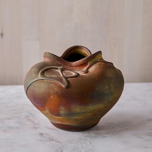 May include: A handcrafted ceramic vase with a unique, organic shape. The vase features a blend of earthy tones, including brown, orange, and green, with a metallic sheen. An abstract design adorns the side. This decorative piece is ideal for home decor.