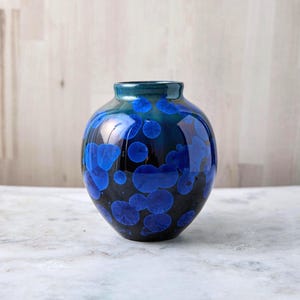 May include: A small, round ceramic vase with a dark blue and black glaze. The vase features a pattern of blue crystalline formations. The top rim is a darker shade of blue. The vase is displayed on a marble surface.