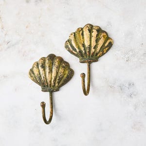 May include: Two antique-style seashell wall hooks. The hooks are made of metal with a distressed gold and green finish. The seashell design is detailed, and the hooks are curved.
