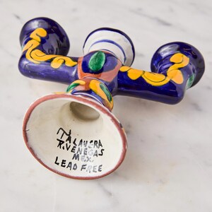 May include: A Talavera ceramic candle holder with three arms. The candle holder is painted with a dark blue base color and decorated with yellow and orange floral designs. The base is white with black text that reads "Talavera R. Venegas Mex. Lead Free."
