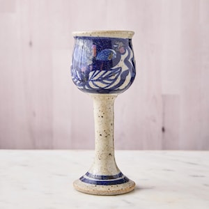 May include: A handmade ceramic wine glass with a speckled off-white stem and base. The bowl is a deep blue with white leaf and floral designs. The rim is off-white with blue accents. A unique piece for serving wine.