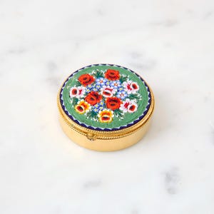 May include: A round, gold-colored trinket box with a mosaic floral design on the lid. The mosaic features red, white, and yellow flowers on a green background, framed by a blue border. The box is approximately 2 inches in diameter.
