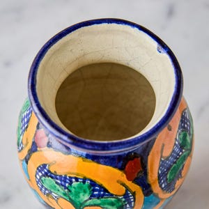 May include: A hand-painted ceramic vase with a wide opening and a vibrant, colorful design. The vase features a blue rim, with orange, green, and blue floral patterns. The interior is a light beige color.