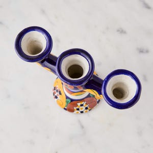 May include: A hand-painted ceramic candelabra with three openings for candles. The candelabra is primarily dark blue with yellow, orange, and green floral designs. The openings are white with a dark blue trim.