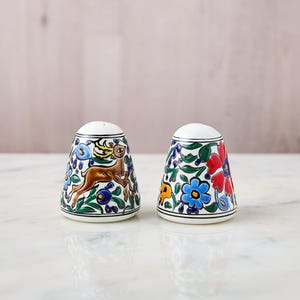 May include: A pair of ceramic salt and pepper shakers. One shaker features a brown deer leaping amidst blue flowers and green leaves. The other shaker is decorated with red and blue flowers and green leaves. Both have white tops.