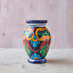 May include: A hand-painted ceramic vase with a dark blue base and rim. The vase features a colorful floral design with orange, green, and blue accents. The vase is approximately 7 inches tall and sits on a white marble surface.