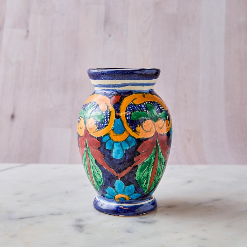 May include: A hand-painted ceramic vase with a dark blue base and rim. The vase features a colorful floral design with orange, green, and blue accents. The vase is approximately 7 inches tall and sits on a white marble surface.