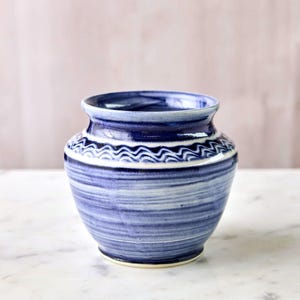 May include: A blue and white ceramic vase with a rounded body and a slightly flared rim. The vase features horizontal stripes and a decorative wave pattern around the neck. The color palette includes shades of blue and white.
