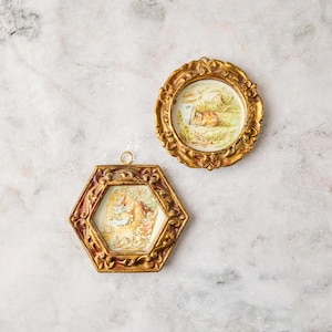 May include: Two decorative wall ornaments featuring ornate gold-toned frames. One is a circular frame containing an illustration of a small brown mouse in grass, and the other is a hexagonal frame displaying a scene with figures in a garden setting.