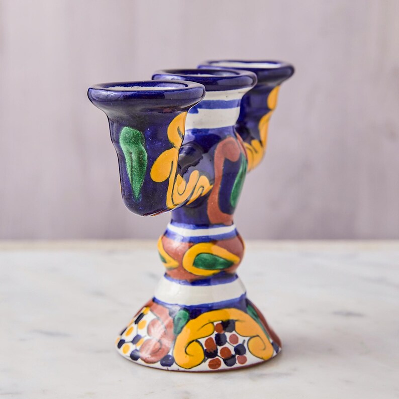 May include: A hand-painted ceramic candle holder with three openings. The holder features a dark blue base with colorful floral and abstract designs in yellow, green, and brown. White stripes accent the neck of the holder.