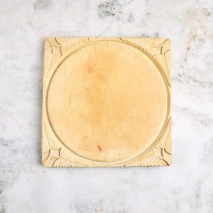 May include: A square, light-colored wooden cutting board with a circular center. The corners feature carved decorative elements. The board is approximately 12 inches wide and 12 inches long.