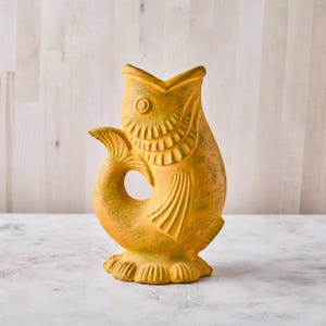 May include: A ceramic fish-shaped vase in shades of yellow and green. The vase has an open mouth and a tail fin. The fish has detailed scales and fins, and sits on a scalloped base. The vase is a decorative item.