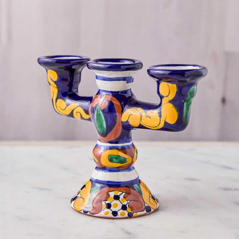 May include: A hand-painted ceramic candelabra with three candle holders. The candelabra is primarily dark blue with yellow, green, and red floral designs. White and yellow stripes accent the base and candle holders. The base is approximately 5 inches wide.