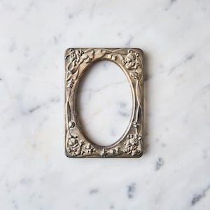 May include: A rectangular silver-toned picture frame with an oval opening. The frame is decorated with embossed cartoon characters and floral designs. The frame is placed on a white marble surface.