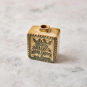 May include: A small, rectangular ceramic vase with a speckled beige finish. The front features a recessed square panel with a dark green relief design of two birds perched on a tree. The edges of the panel have a decorative sawtooth pattern.