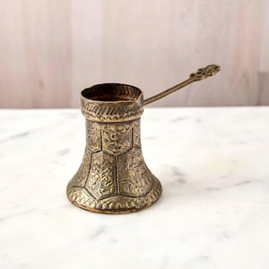 May include: A vintage brass coffee pot with an ornate floral design. The pot has a long handle and a wide base, tapering to a narrower top. The surface is covered in detailed engravings, giving it a classic, antique look.