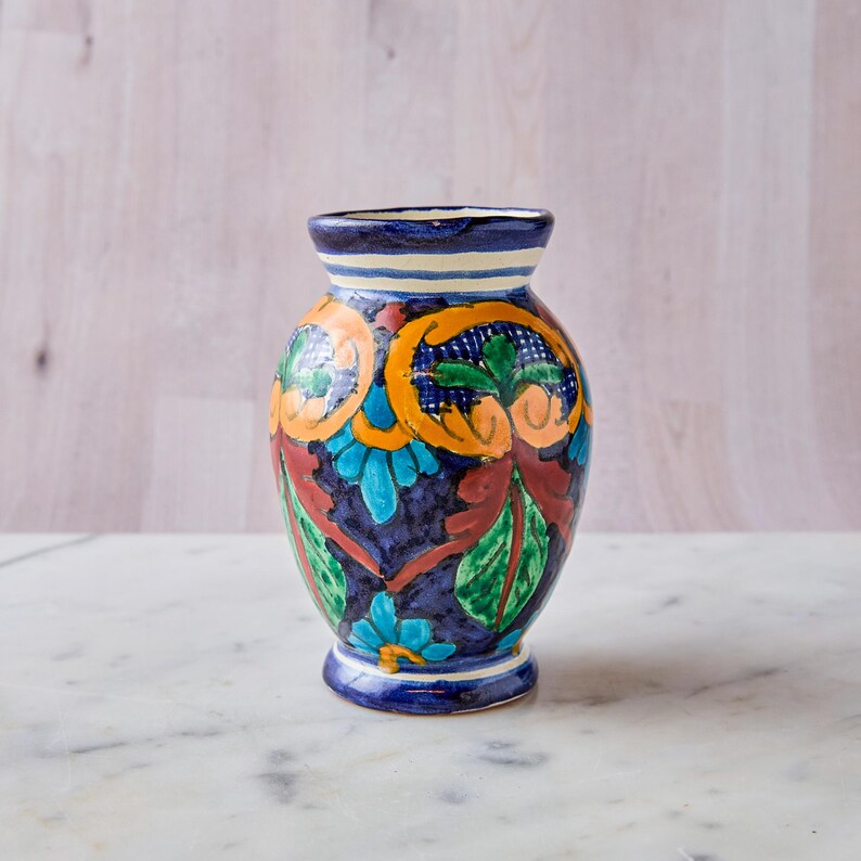 May include: A hand-painted ceramic vase with a dark blue base and a flared rim. The vase features colorful floral designs in orange, teal, and green against a dark blue background. The vase is approximately 6 inches tall.