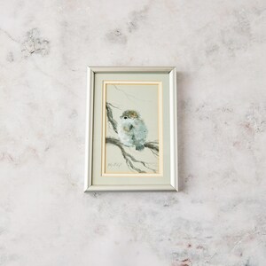 May include: A framed watercolor painting featuring a small, fluffy bird perched on a thin, dark branch. The artwork is set within a light-colored mat and a metallic silver frame, showcasing soft gray and brown tones against a pale background.