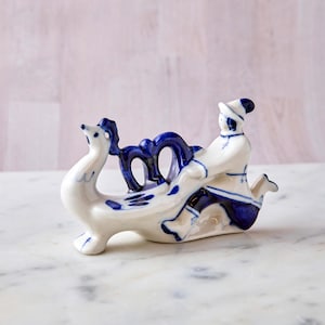 May include: A white and blue ceramic figurine depicting a person riding a bird-like creature. The figurine has intricate blue detailing and is set against a soft pink background. The style is reminiscent of traditional folk art.