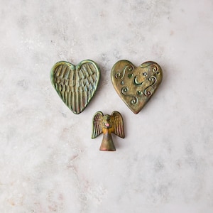 May include: Three decorative ceramic pieces with a metallic bronze and green finish. One heart-shaped piece features embossed angel wings, another heart-shaped piece displays a crescent moon and stars, and a small angel figurine stands below.