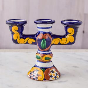 May include: A hand-painted ceramic candelabra with three candle holders. The candelabra is primarily dark blue with yellow, orange, and green floral designs. White stripes accent the base and the candle holders. The base is approximately 6 inches wide.