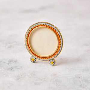 May include: A circular picture frame with a gold-toned metal border featuring an intricate mosaic inlay of bright orange and light blue tiles. The frame stands on two small, matching mosaic-decorated feet and contains a plain, cream-colored center.