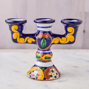 May include: A hand-painted, three-armed ceramic candle holder. The candle holder is primarily dark blue with yellow, orange, and green floral designs. The base and arms have decorative patterns, and the candle cups are white.