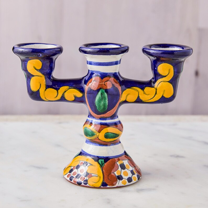 May include: A hand-painted, three-armed ceramic candle holder. The candle holder is primarily dark blue with yellow, orange, and green floral designs. The base and arms have decorative patterns, and the candle cups are white.