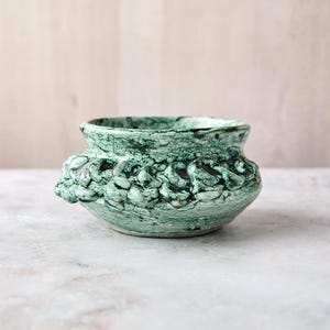 May include: A small, round ceramic bowl featuring a mottled green and white glaze with a crackled texture. The exterior is decorated with a series of raised, interconnected loop-like shapes encircling the upper portion of the vessel.