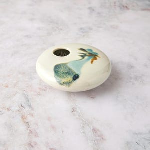 May include: A low, disc-shaped ceramic flower frog vase with a glossy off-white glaze. The top surface features a circular opening containing a metal pin holder, accented by a decorative abstract splash of blue, green, and brown glaze.