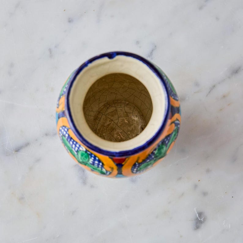 May include: A round, hand-painted ceramic container with a wide opening. The exterior features a blue background with orange, green, and red decorative patterns. The rim is blue, and the interior is beige with a cracked texture.