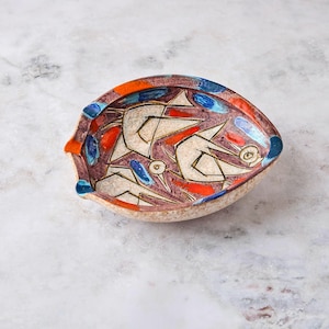 May include: A shallow, asymmetrical ceramic bowl featuring a speckled beige base with a hand-painted design of two stylized birds in black outlines. The interior is decorated with vibrant orange and blue accents against a deep purple background.