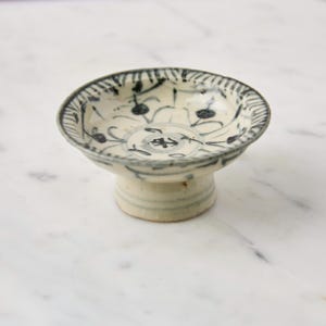 May include: A small, off-white ceramic bowl with a pedestal base. The bowl features a blue and white floral pattern around the rim and inside. The pedestal base is a light beige color. The bowl is likely vintage or antique.