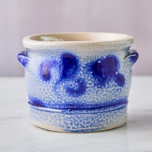 May include: A white ceramic crock with blue glaze and two handles. The crock has a speckled texture and a blue glaze that is unevenly applied.