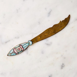 May include: An antique butter knife with a brass blade and a decorative handle. The handle is adorned with a mosaic pattern in shades of red, blue, and white. The blade has a unique, curved shape.