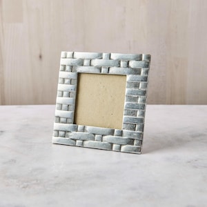 May include: A square picture frame with a metallic silver finish and a textured, woven basket-weave pattern across the border. The frame features a plain, light beige insert in the center, providing a neutral display area for a photograph.