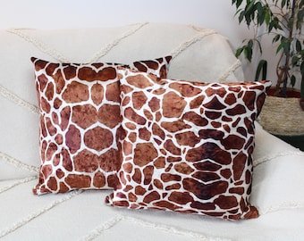 May include: Two square throw pillows with a giraffe print pattern in shades of brown and white. The pillows are placed on a cream-colored sofa. The pillows have a soft, textured appearance and add a touch of safari style to the decor.