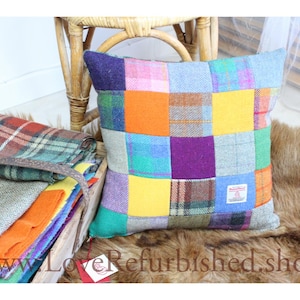 May include: A colourful patchwork cushion with squares of purple, orange, yellow, and green. A stack of blankets and a wooden chair are in the background. The cushion is on a brown fur-like surface.