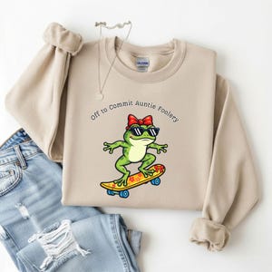 May include: Beige sweatshirt with the text "Off to Commit Auntie Foolery" and a graphic of a green frog wearing sunglasses and a red bow riding a skateboard. The skateboard is yellow with a floral design.