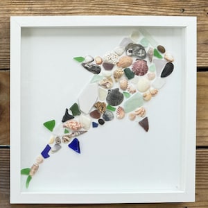 May include: A framed artwork featuring a shark design made from seashells and sea glass. The shark is composed of various shells, glass pieces in green, blue, and brown, and is set against a white background. The frame is white.