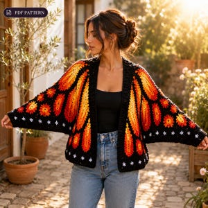 Puede incluir: Cárdigan negro de ganchillo con un diseño de alas de mariposa en tonos naranja y amarillo. El cárdigan tiene una base negra con detalles de estrellas blancas y el texto "PDF PATTERN". Una prenda única, hecha a mano.