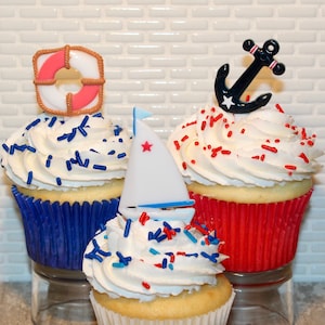 May include: Three cupcakes decorated with red, white, and blue sprinkles. The cupcakes are topped with a life preserver, an anchor, and a sailboat.