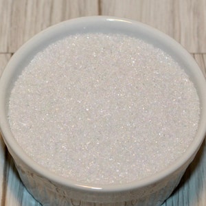 May include: A white, ceramic bowl filled with white, sparkling granules. The granules have a shimmering, iridescent quality, catching the light. The bowl is round with a slightly flared rim. The background is a light wood grain.