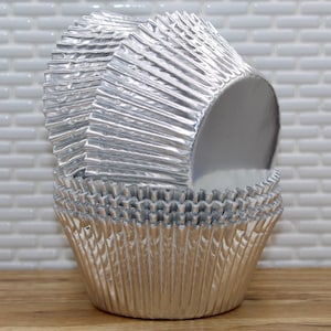 May include: A stack of silver cupcake liners, made of foil, with a fluted design. The liners are stacked in a pyramid shape, with the top liner being the largest.