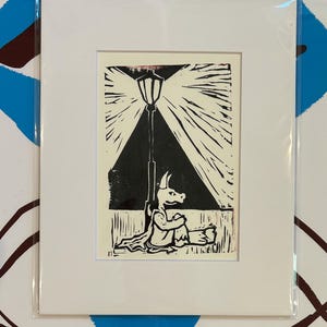 May include: A black and white linocut print of a creature sitting under a streetlight. The creature has a long snout and is wearing a cloak. The streetlight is casting a beam of light down on the creature.